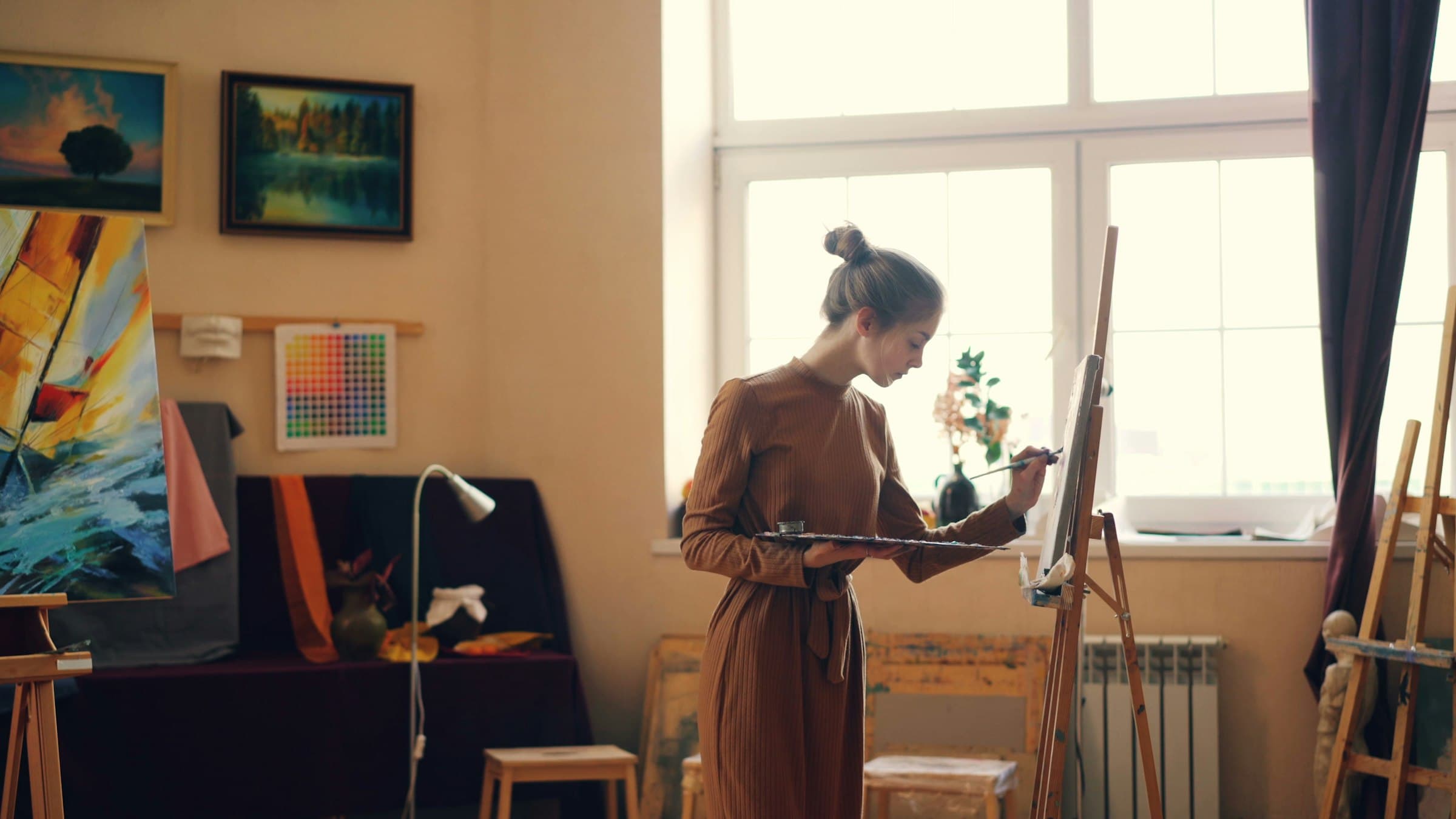 Painter at work in a bright studio mixing colors on a palette beside a canvas